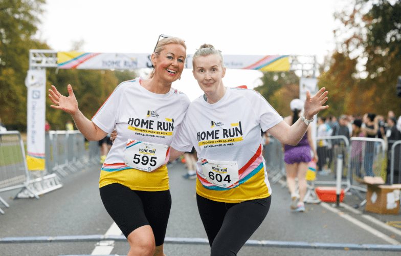 Alt text: Two participants wearing numbered vests stand side by side on a road at a race finish line with arms raised outward. Both wear white “Simon Home Run” event T‑shirts with multicolours. A branded finish gantry, metal crowd barriers, and other participants walking on the course are visible in the background, with trees lining the route.